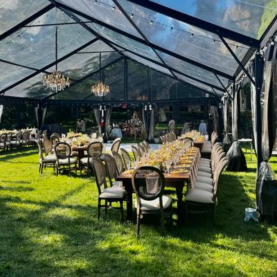 Wedding reception setup with elegantly decorated tables on the grass under a clear plastic tent.
