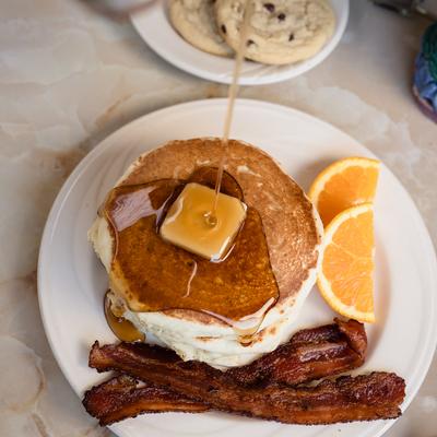 Buttermilk pancakes with butter, bacon, and orange slices, drizzled with syrup.