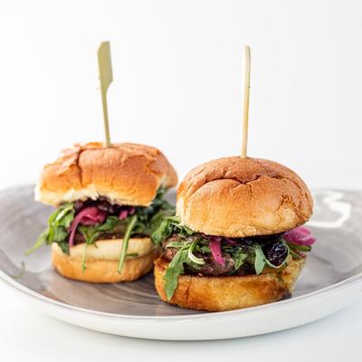 Two burger sliders with arugula and pickled onions sit on a gray plate, white background.