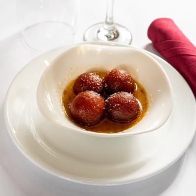 A white bowl of Gulab jamun served on a white plate, set on a white tablecloth.