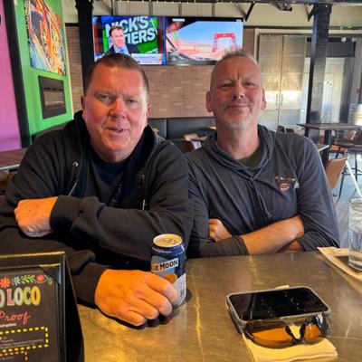 two guys sitting at the bar smiling, one holding a beer can..