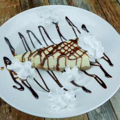 New York cheesecake slice drizzled with chocolate on a white plate.