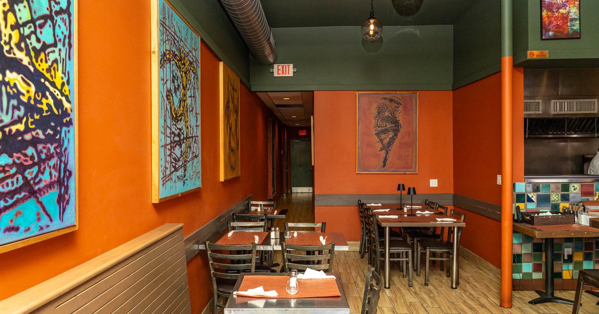 Interior of a restaurant with orange walls, abstract paintings, wooden furniture, and tiled kitchen divider