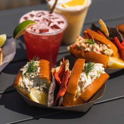 Two lobster rolls with sides of tortilla chips, served alongside couple of cocktails.