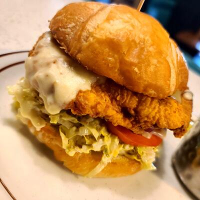 A close-up of a fried chicken Caesar croissant sandwich topped with cheese sauce..
