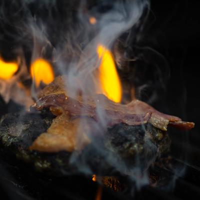 Bacon on a burger grilling over open flames with smoke rising above.