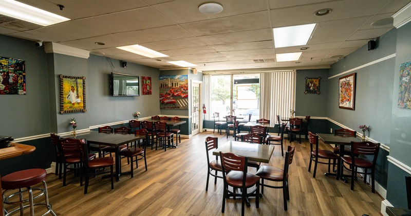 Interior, wooden tables and chairs, colorful paintings on gray walls, and large windows with blinds