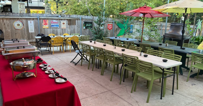 Outdoor patio with long tables and green chairs set for dining, Red tablecloth with buffet dishes