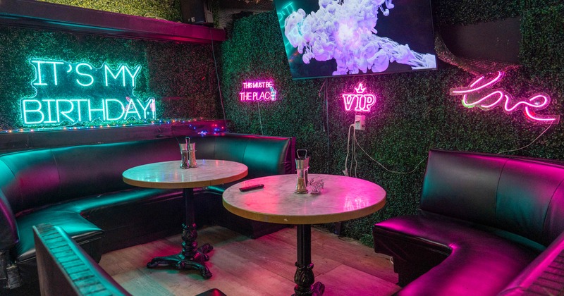 Lounge with green foliage, neon signs, two round tables, black leather booths, and a TV
