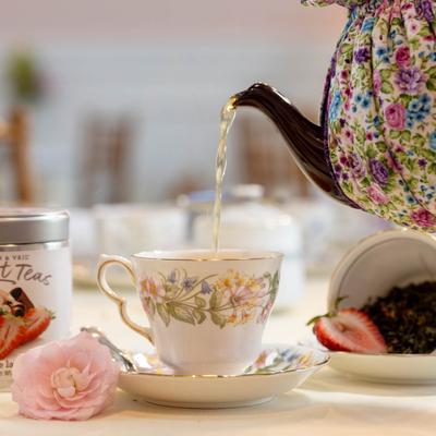 Floral tea cozy–covered pot pouring tea into a cup.