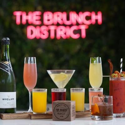 Variety of colorful cocktails and a champagne bottle sit under a neon 