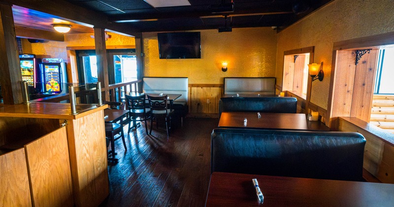 Wood-paneled restaurant or bar interior with booths, tables, a wall-mounted TV, gaming machines, and warm amber lighting