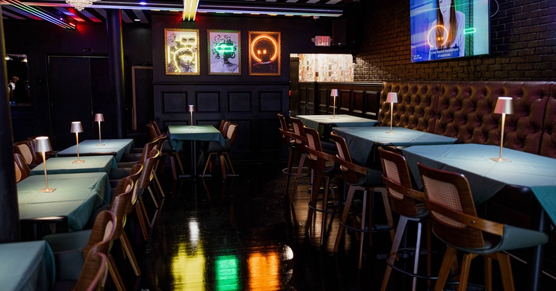 Interior of a stylish bar with teal-covered tables, wooden chairs, neon lights, and a flat-screen TV