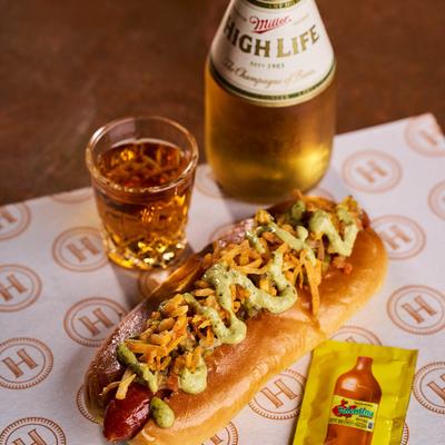 A gourmet hot dog, a bottle of beer, a shot glass filled with a beverage, and a packet of hot sauce.