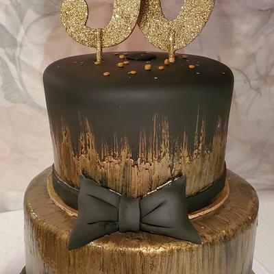 A cake with black frosting and gold brushstrokes, gold 60 topper, and a black bow.