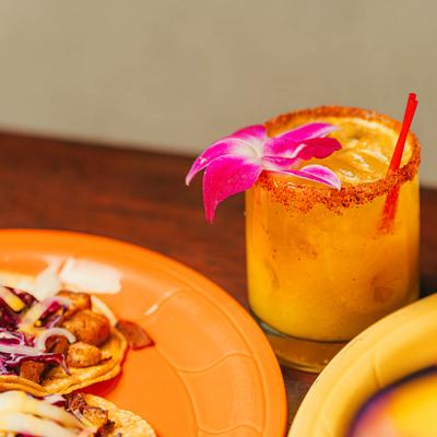Chili-rimmed mango margarita with orchid garnish beside a plate of tacos.
