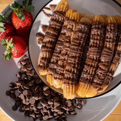 Plate of churros drizzled with chocolate, surrounded by strawberries and chocolate chips.