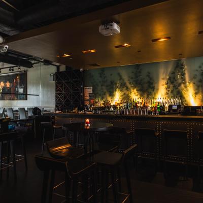 Dimly lit bar with illuminated bottles on a wide counter, surrounded by tall chairs.