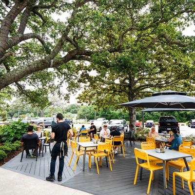 People sitting on outdoor patio with yellow chairs and gray tables under large trees.
