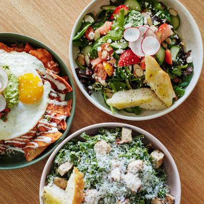 Bowls of chilaquiles with fried eggs, kale Caesar salad, and farmer's salad on a table.