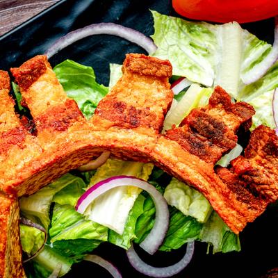 Crispy fried chicharron sits atop fresh lettuce, red onion slices, and a tomato on a dark plat.