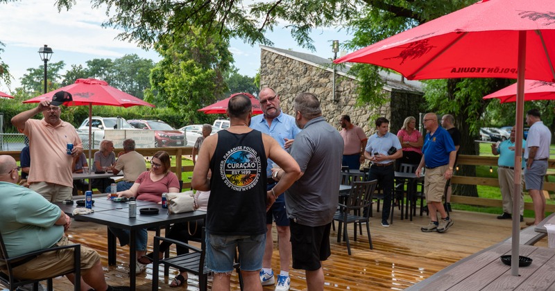 People gather on an outdoor wooden patio with tables and red umbrellas