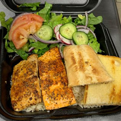 Grilled salmon on rice with toasted bread and a fresh salad in a black takeout container.