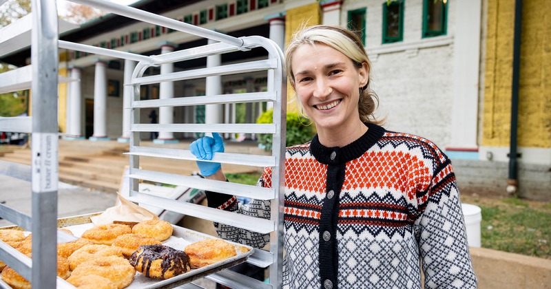 Outdoors, a smiling employee stands by a tray of fresh pastries on a bakery rack