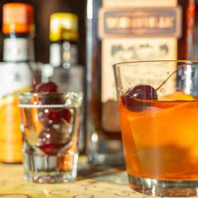 Old Fashioned cocktail with cherries, blurred bottles in the background.
