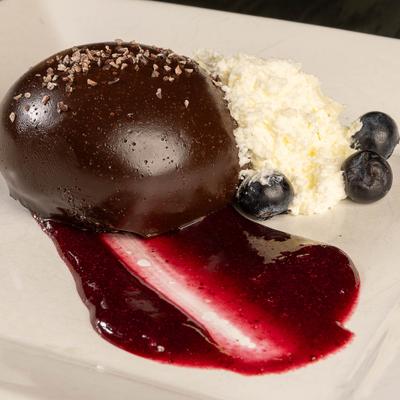 A glossy chocolate dome on a white plate with raspberry sauce, whipped cream and blueberries.