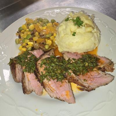 Sliced meat topped with herbs, served with mashed potatoes and vegetables.
