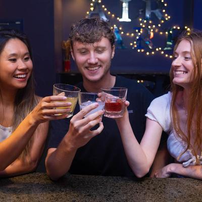 Three people toasting with drinks at a bar with festive lights in the background.
