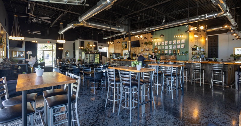 Interior of a restaurant featuring tall tables and chairs, a bar area with shelves of bottles
