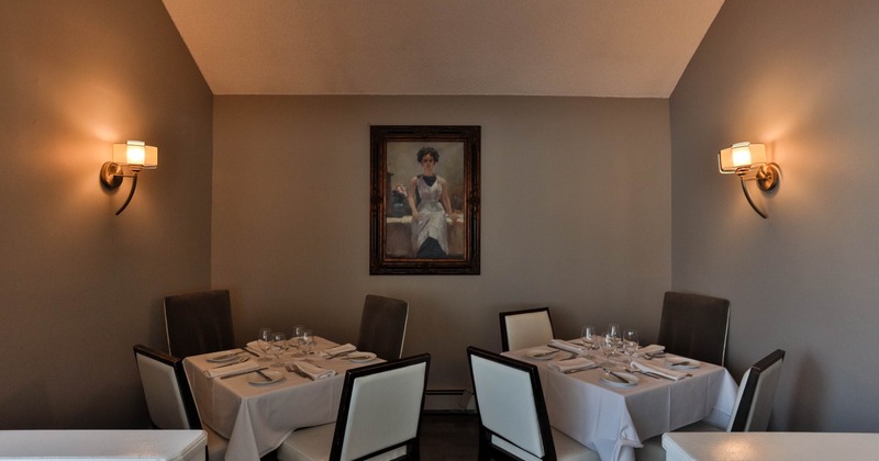 A small dining area with two set tables, cushioned chairs, a framed painting
