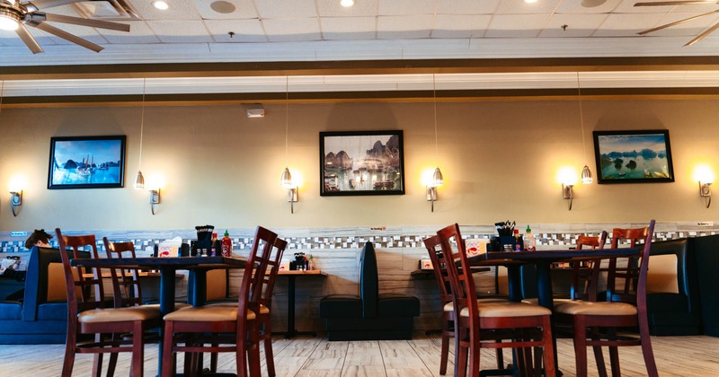 Interior of a restaurant with round wooden tables, brown-cushioned chairs, framed landscape pictures