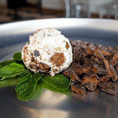A scoop of ice cream on mint leaves next to dark chocolate chunks, set on a metal plate.
