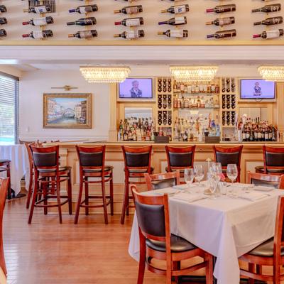 Bright restaurant interior with bar seating, white-tablecloth tables, and wall wine display.