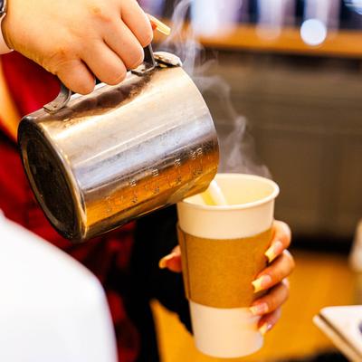 A barista pouring steaming milk from a metal pitcher into a paper cup with a sleeve.