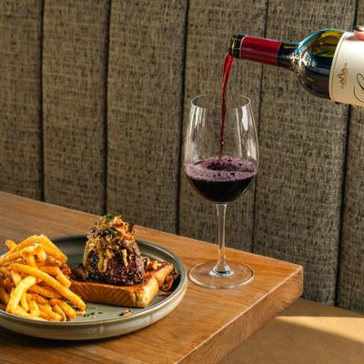 Red wine being poured into a glass, alogside a steak sandwich with fries.