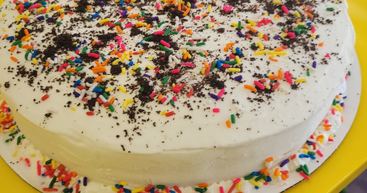 White frosted cake topped with rainbow sprinkles and chocolate drizzle