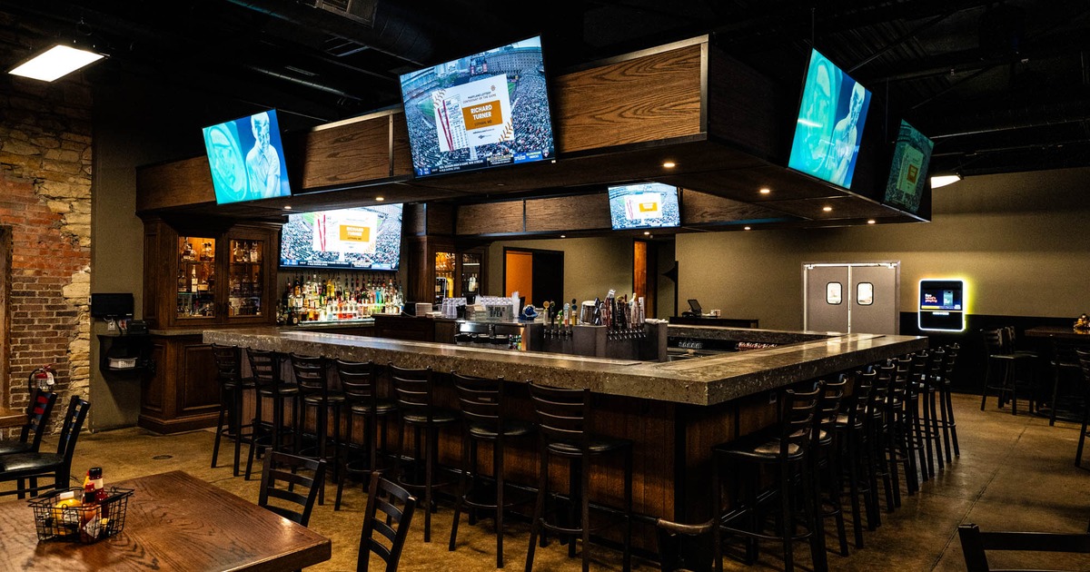 ull indoor sports bar with multiple large screens, a long bar with stools, and tables