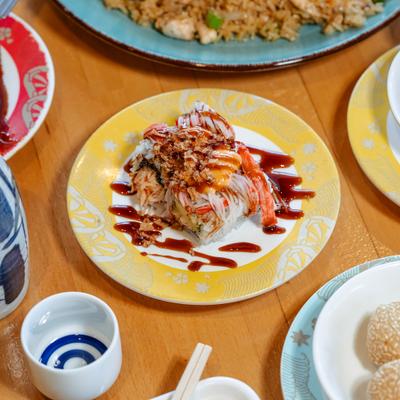 Fiesta roll sushi with sauce drizzle on the plate, served on a wooden table with assorted foods.