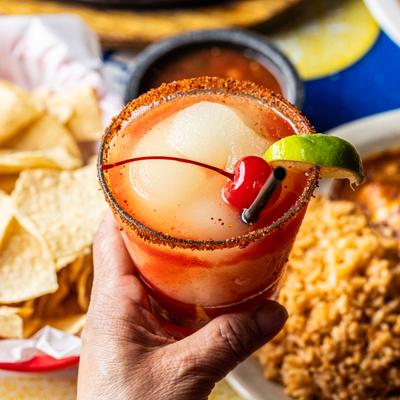 A hand holding a glass of Frozen Strawberry Margarita, with food plates in the background.