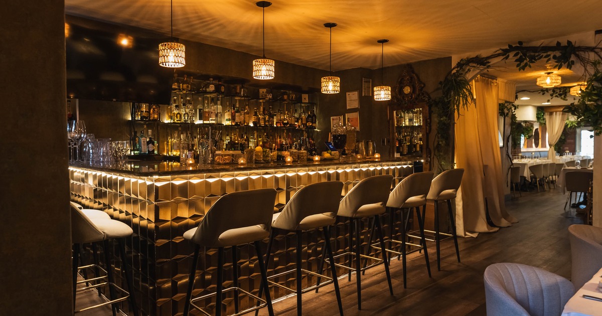 A warmly lit restaurant bar with patterned decor, bar stools, and shelving stocked with bottles