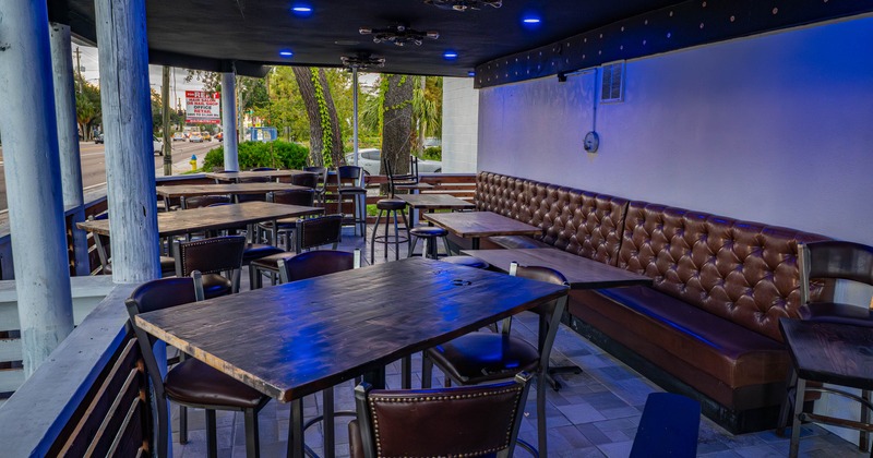 Outdoor seating area, wooden tables, leather chairs, tufted bench under blue lights