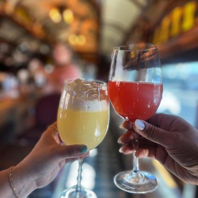 Two hands holding drinks in a bar, with a yellow cocktail and a red beverage.