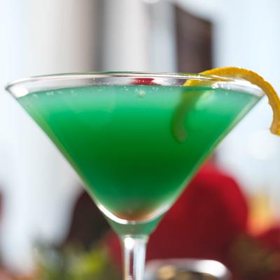 Green cocktail served in martini glass, garnished with orange peel and a cherry.
