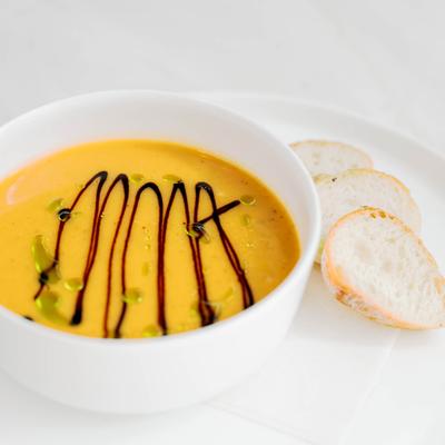 Bowl of orange soup with balsamic glaze and green oil on a white plate with bread slices.
