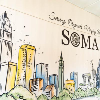 Colorful cityscape mural with the SOMA logo and handwritten slogan.