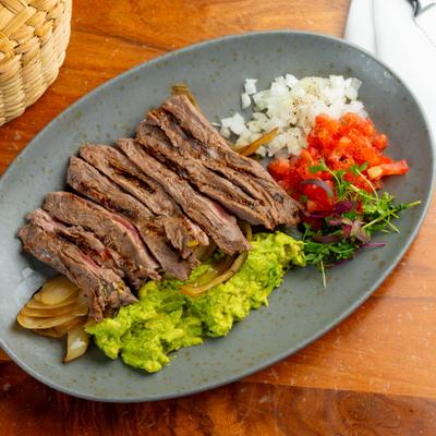 Sliced steak served alongside guacamole, diced tomatoes, onions, and microgreens.